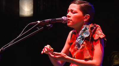  En el escenario, Natalia, con el cabello recogido y ojos cerrados, canta ante el micrófono. Las puntas de sus dedos están a punto de tocarse. La cantante lleva un vestido rojo con grandes flores (una azul, las otras rojas) bordadas a la altura del pecho y en el hombro izquierdo.