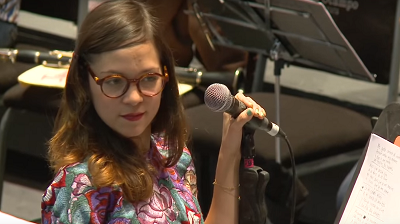 Durante el ensayo, Natalia toma el micrófono con su mano izquierda y voltea hacia su derecha. Lleva una blusa color turquesa con flores lilas bordadas. 