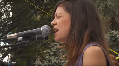  Foto de Natalia cantando en una plaza. Ella está de perfil ante el micrófono, lleva cabello suelto y una blusa morada. 